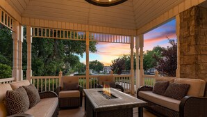 Une terrasse couverte, aménagée avec des meubles en osier, un foyer et offrant une vue sur les arbres et le coucher de soleil.