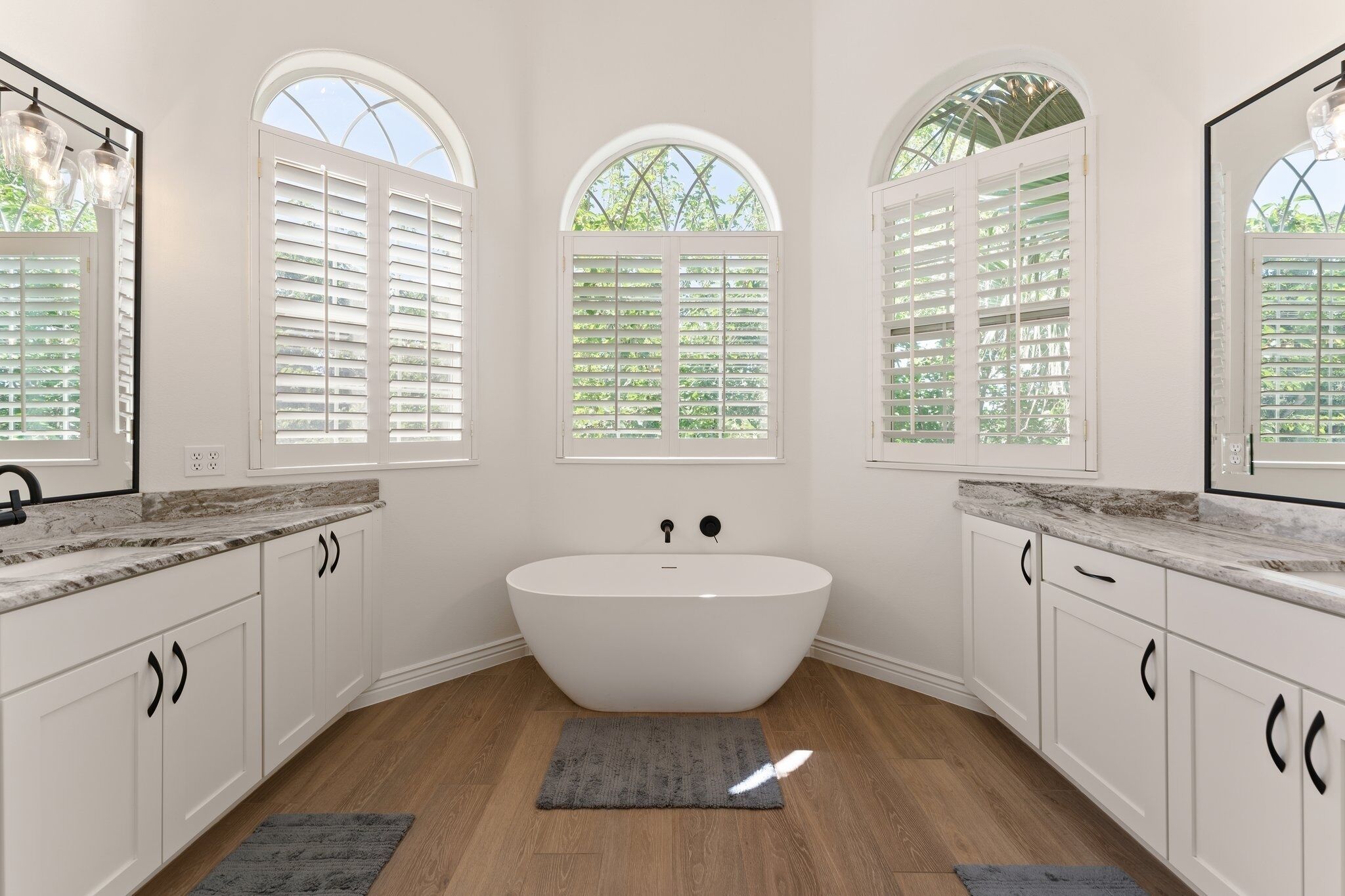 A bathroom with a freestanding bathtub, white cabinetry, and large arched windows.