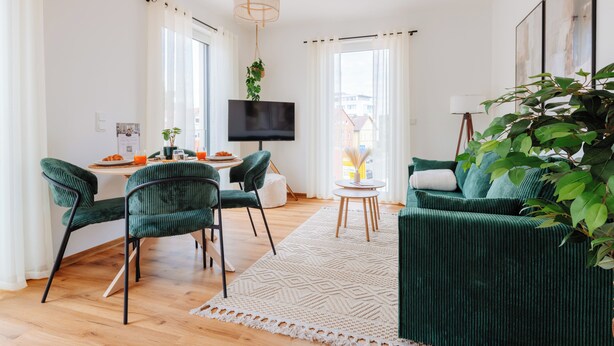 Una sala de estar moderna con un sofá verde, una mesa de comedor con sillas, un televisor de pantalla plana y un ventanal con cortinas.