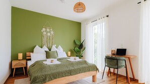 Un dormitorio con una pared verde, una cama de madera, almohadas blancas, una mesita de noche con una lámpara, un escritorio con una silla y un ventanal con cortinas.