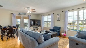 Una sala de estar amplia con sofá, mesa de centro y televisor. Hay cortinas, un ventilador de techo y un letrero que dice "SEAS THE DAY".