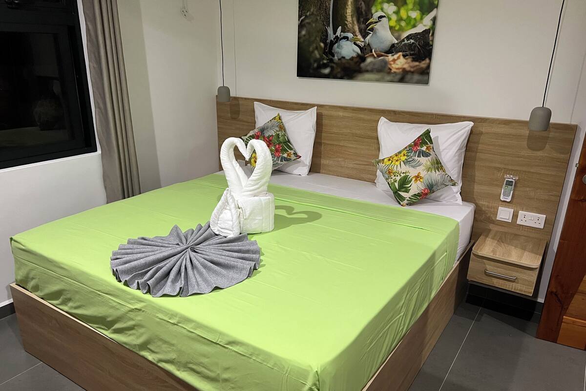 A neatly made bed with a green bedspread, two pillows, and a towel shaped like swans.