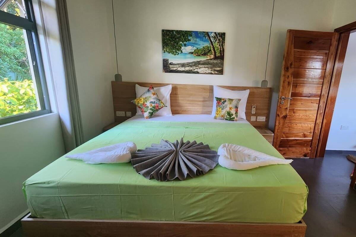 A neatly made bed with green bedding, a tropical painting on the wall, and a wooden door.