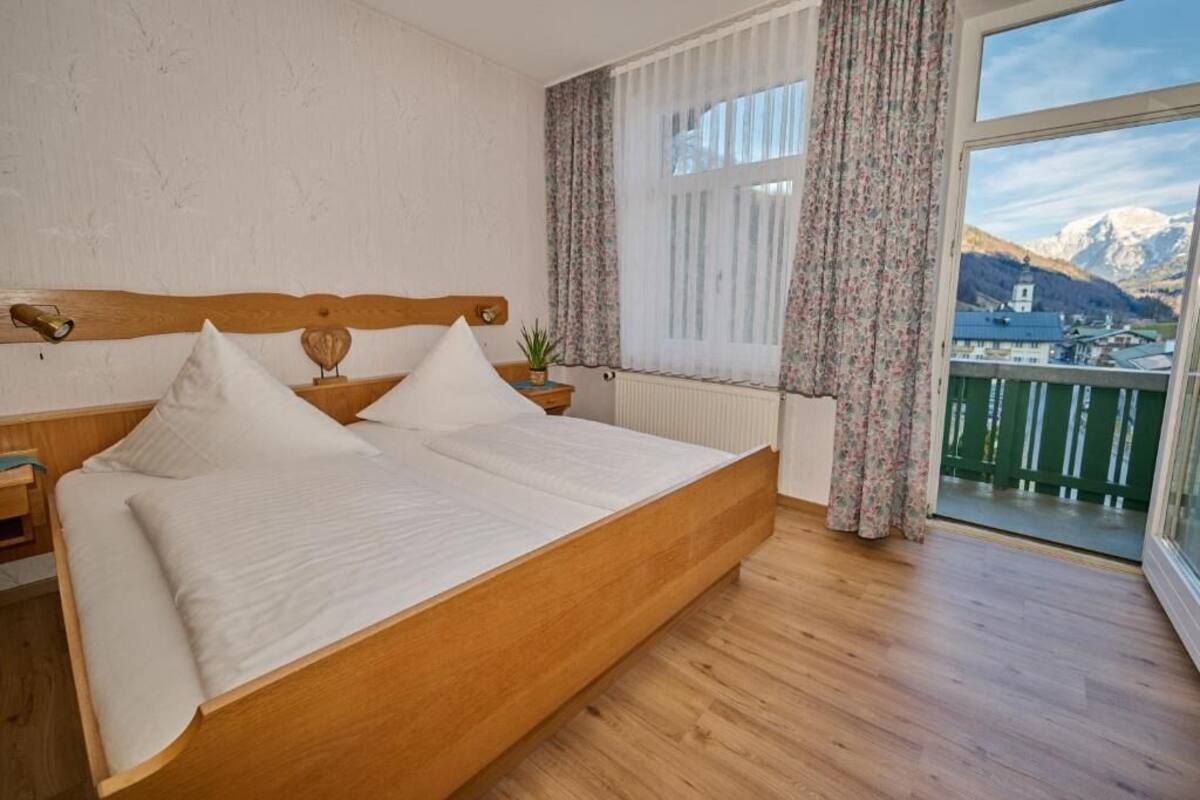 A bedroom with a wooden bed, white bedding, a nightstand, a bedside table, a window with curtains, and a view of mountains.