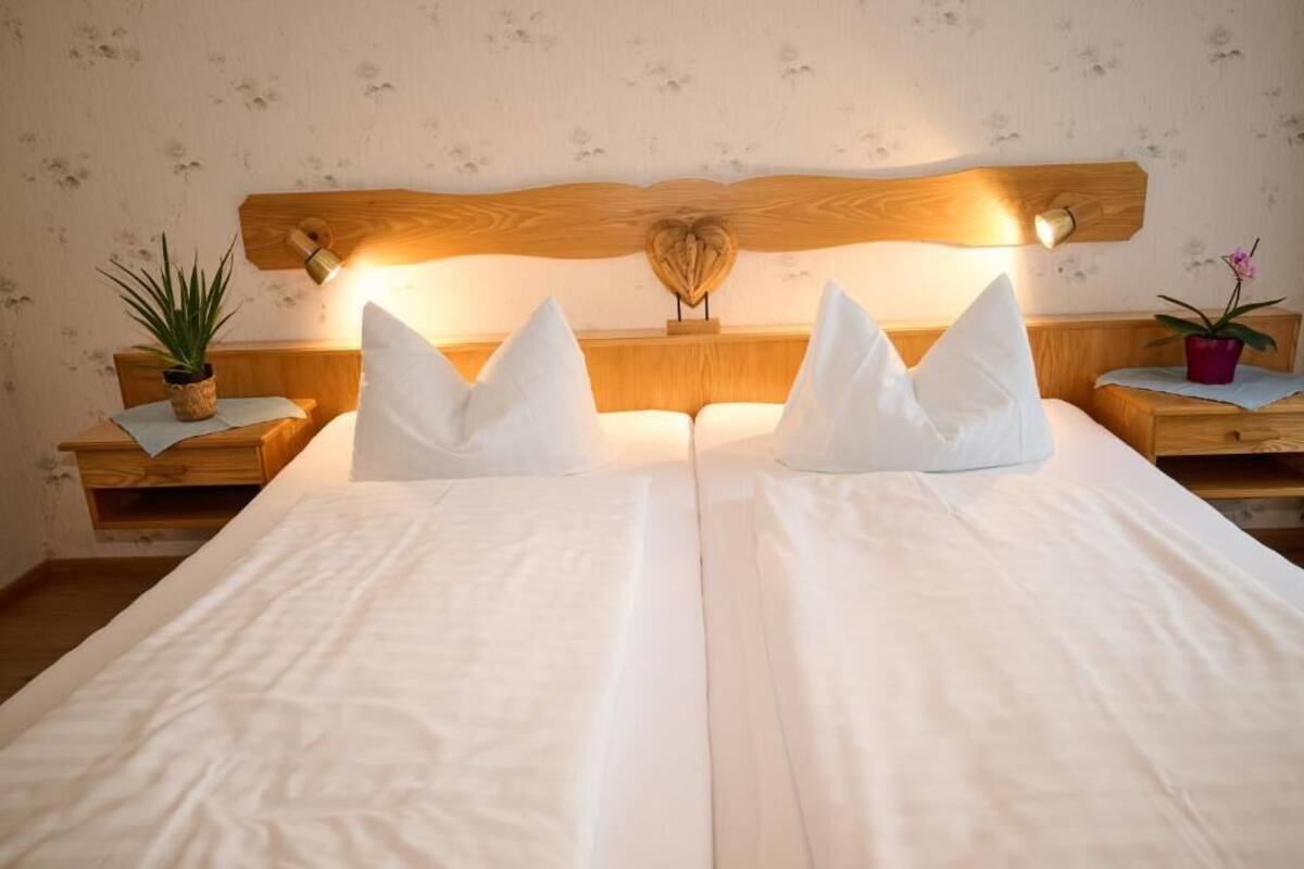 A double bed with white bedding and two pillows, wooden headboard with a heart-shaped design, and two bedside tables with potted plants.
