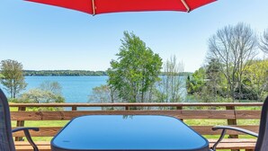 Een houten terras met een tafel en stoelen onder een rode parasol, met uitzicht op een meer, bomen en een verre oever.