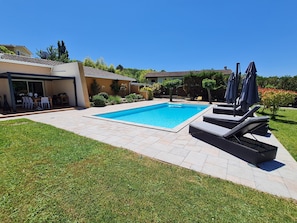 Piscina retangular com deck ladrilhado, cercada por grama e algumas árvores.