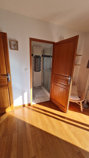 Quarto com piso de madeira, porta para banheiro com chuveiro e uma cadeira.