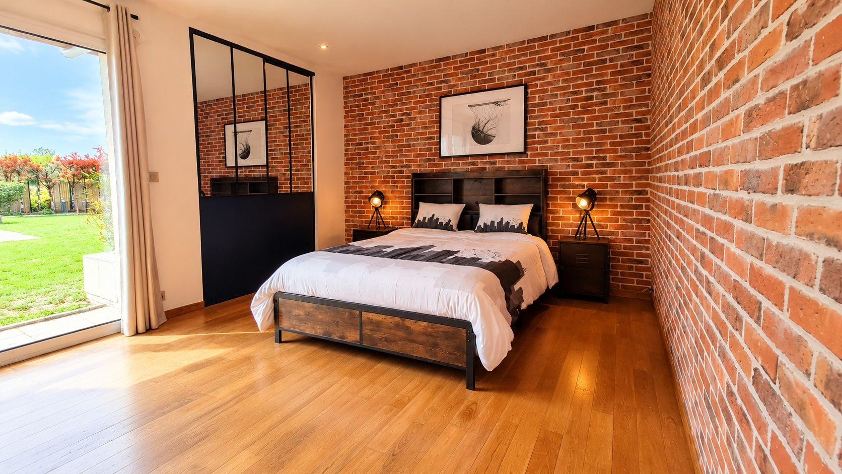 Une chambre avec un mur en briques, un plancher en bois, un lit avec une tête de lit, deux tables de chevet et une grande fenêtre.