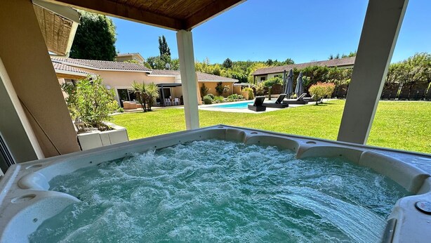Ein Whirlpool mit Blick auf einen Garten, der einen Pool, Liegestühle und eine überdachte Terrasse bietet.