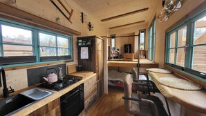 A compact kitchen with a gas stove, sink, and wooden cabinets. There is a dining area with a bench and a chair. Large windows provide natural light and views of the outdoors. - Tiny House Design in a Vacation Park Near an Eco-Village (Steyerberg)