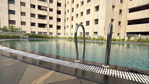 A swimming pool with a metal ladder, surrounded by a tiled deck. In the background, there is a tall building with multiple balconies. - 2-bedroom apartment in delightful Manipal with AC, WiFi, fitness room (Manipal)