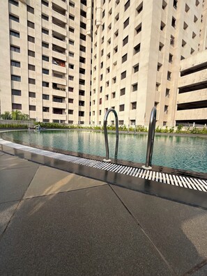 A swimming pool with a metal ladder, surrounded by a tiled deck. In the background, there is a tall building with multiple balconies. - 2-bedroom apartment in delightful Manipal with AC, WiFi, fitness room (Manipal)