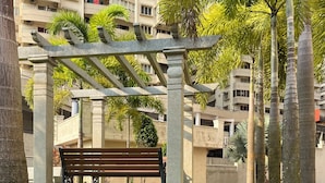 A park area with a wooden bench under a pergola, surrounded by palm trees and a building in the background. - 2-bedroom apartment in delightful Manipal with AC, WiFi, fitness room (Manipal)