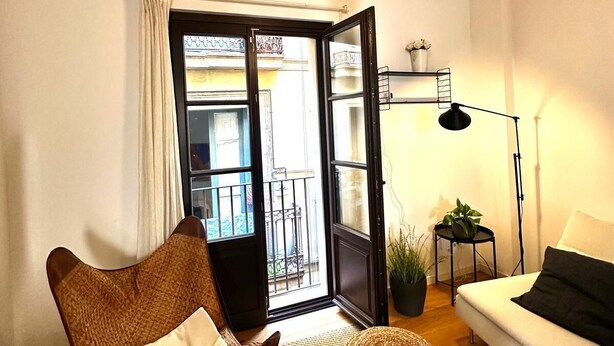 Un accogliente soggiorno con soffitto in legno, un'ampia finestra, un divano bianco, un poggiapiedi intrecciato e una lampada da terra.