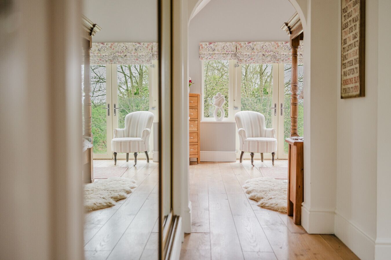 A room with two white upholstered chairs, a wooden cabinet, and a view through large windows.
