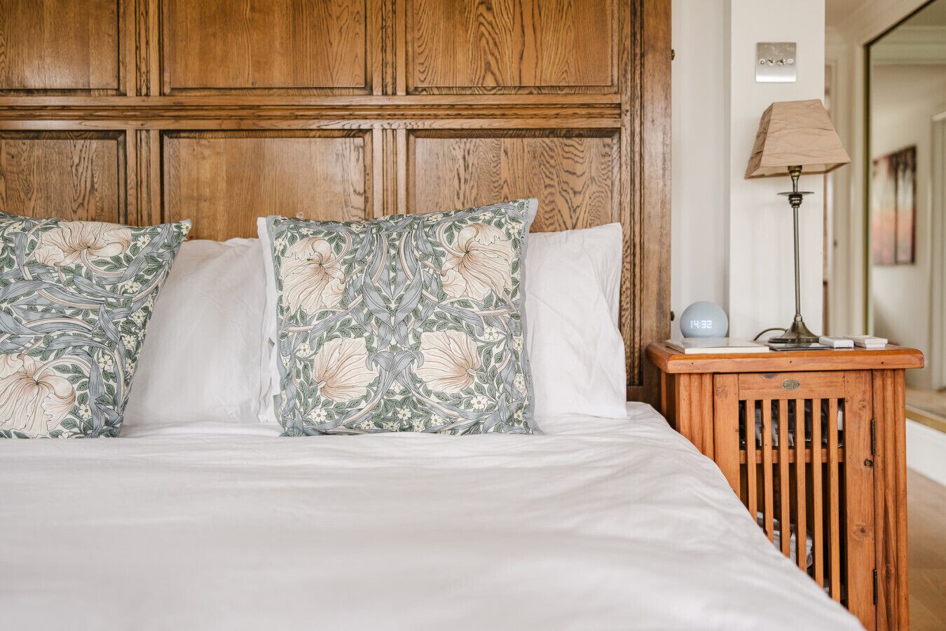 A bed with floral-patterned pillows, a wooden headboard, and a bedside table with a lamp and a clock.