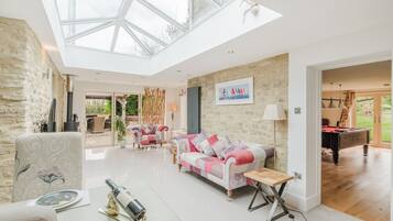 A spacious living room with a high ceiling and skylight, a cozy seating area with a patterned sofa, and a billiards table in the background.