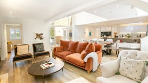Amplia sala de estar con un sofá naranja grande, un sillón blanco y una mesa de centro. Hay un comedor con mesa y sillas, y una cocina con armarios blancos.