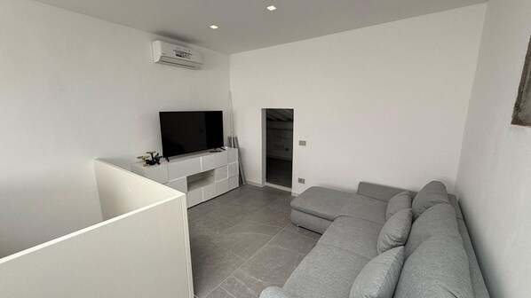 A modern living room with a grey sofa, a flat-screen TV on a white entertainment unit, and a recessed ceiling light.