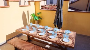 A wooden table set with plates and glasses, a potted plant, and a grill in a sunny outdoor area.