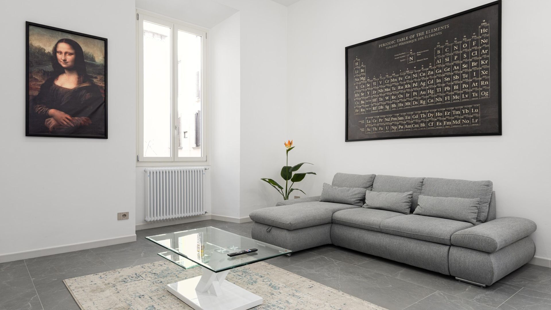 Una sala de estar moderna con un sofá gris, una mesa de centro de vidrio, un cuadro enmarcado y un gran cartel de tabla periódica en la pared.