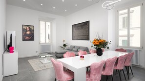 A modern living room with a white sofa, a dining table with pink chairs, and a large window with blinds.
