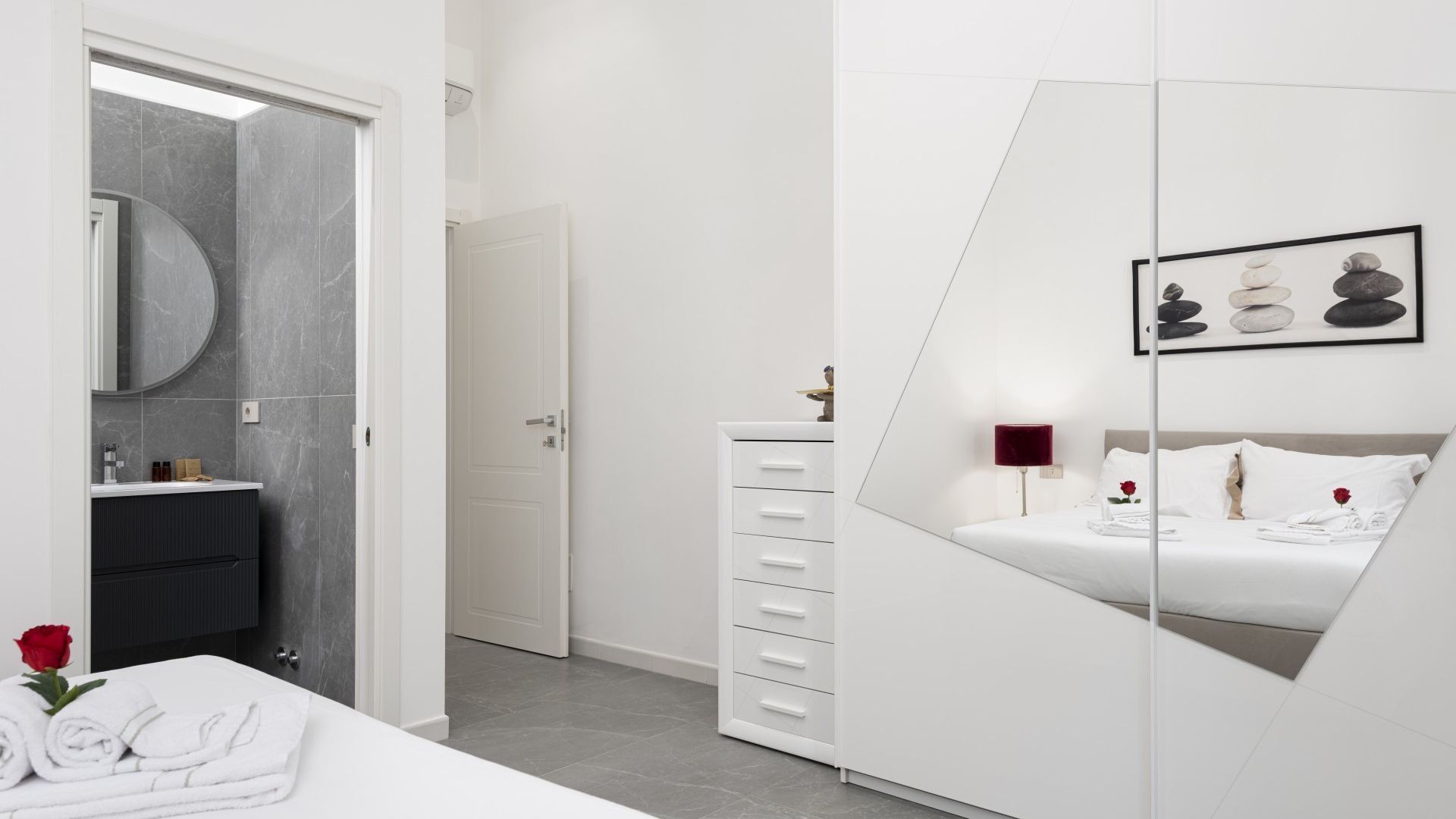 A modern hotel room with a bed, a vanity with a mirror, a dresser, and a framed artwork on the wall.