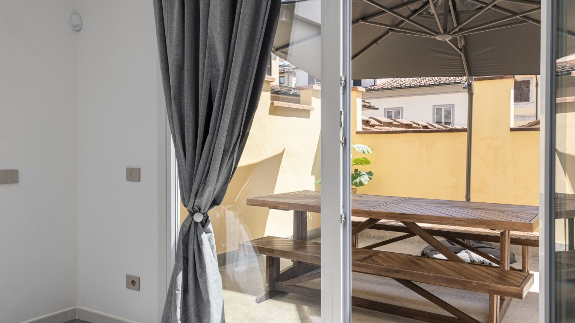 Un balcón con una mesa y sillas de madera, una cortina gris y vistas a edificios al fondo.