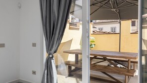 Un balcone con tavolo e sedie in legno, una tenda grigia e vista su edifici sullo sfondo.