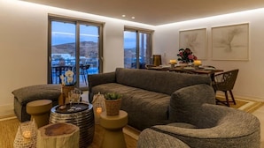 A modern living room with a grey sofa, a round coffee table, and a dining area with a table and chairs. Large windows offer a view of the outdoors.