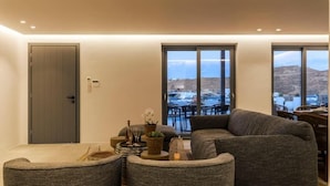 A modern living room with a grey sectional sofa, a small round table with decorative items, and a view of a coastal area through large windows.