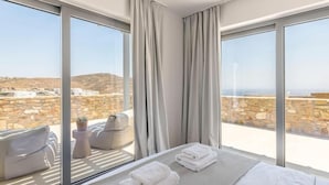 A hotel room with a large bed, two chairs, a small table, and a view of the sea and buildings.
