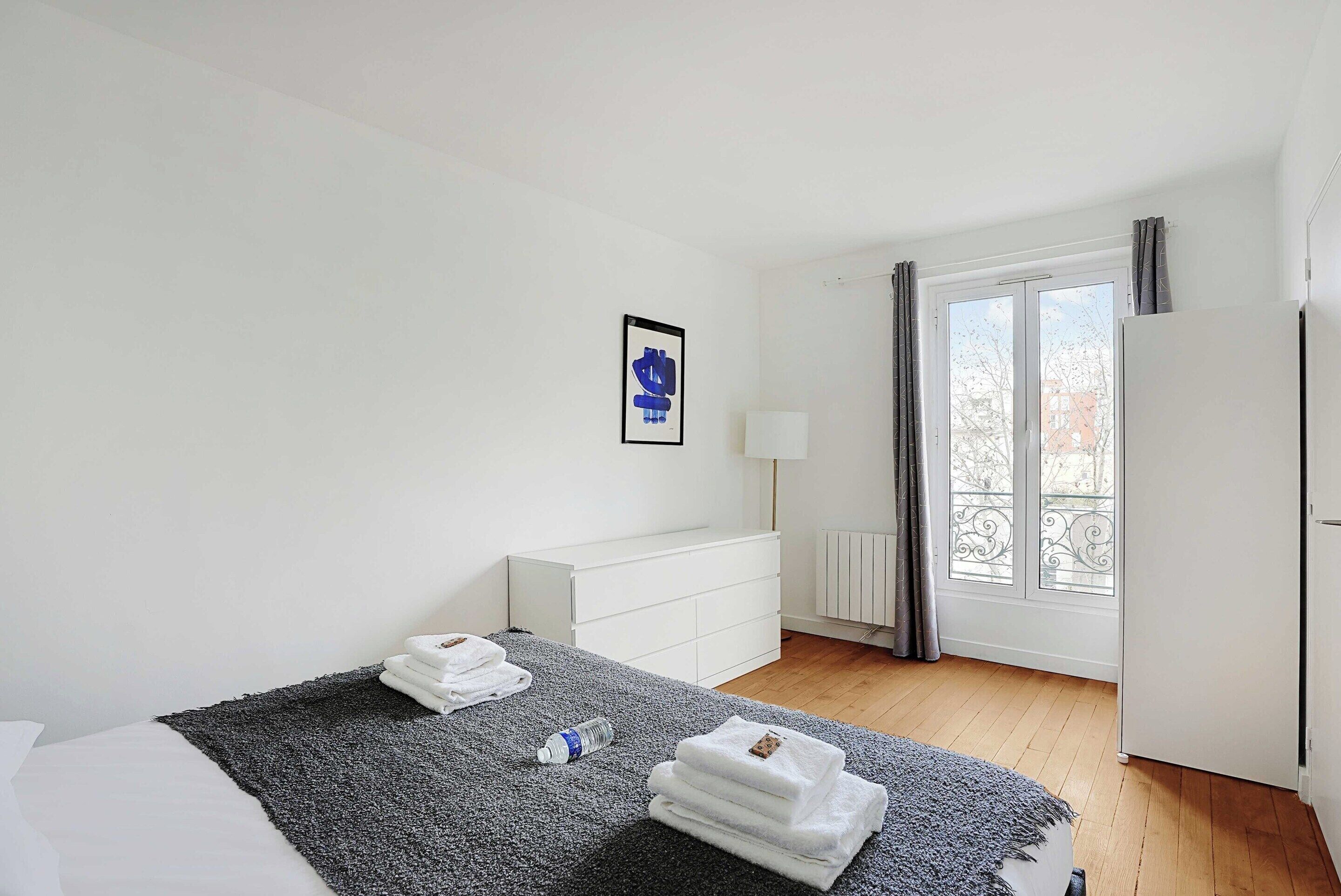 Une chambre à coucher avec un lit, une commode blanche, une fenêtre avec des rideaux et un radiateur.