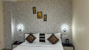 A hotel room with a patterned wallpaper, a bed with white bedding and decorative pillows, two bedside tables with lamps, and framed artwork on the wall. - 45-room hotel in delightful Varanasi with WiFi, AC. Unwind in comfort (Varanasi)