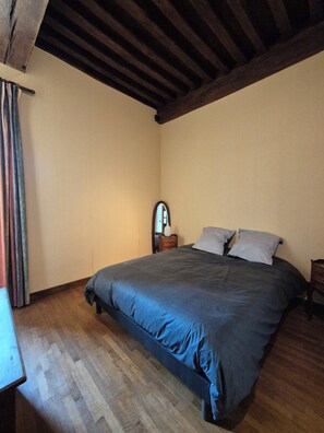A bedroom with a wooden floor, a bed with a dark blue bedspread, a wooden headboard, and a small bedside table with a mirror.