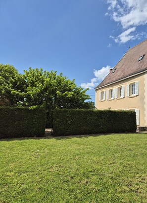 A building with a tiled roof and white shutters, surrounded by a well-maintained lawn and a hedge.