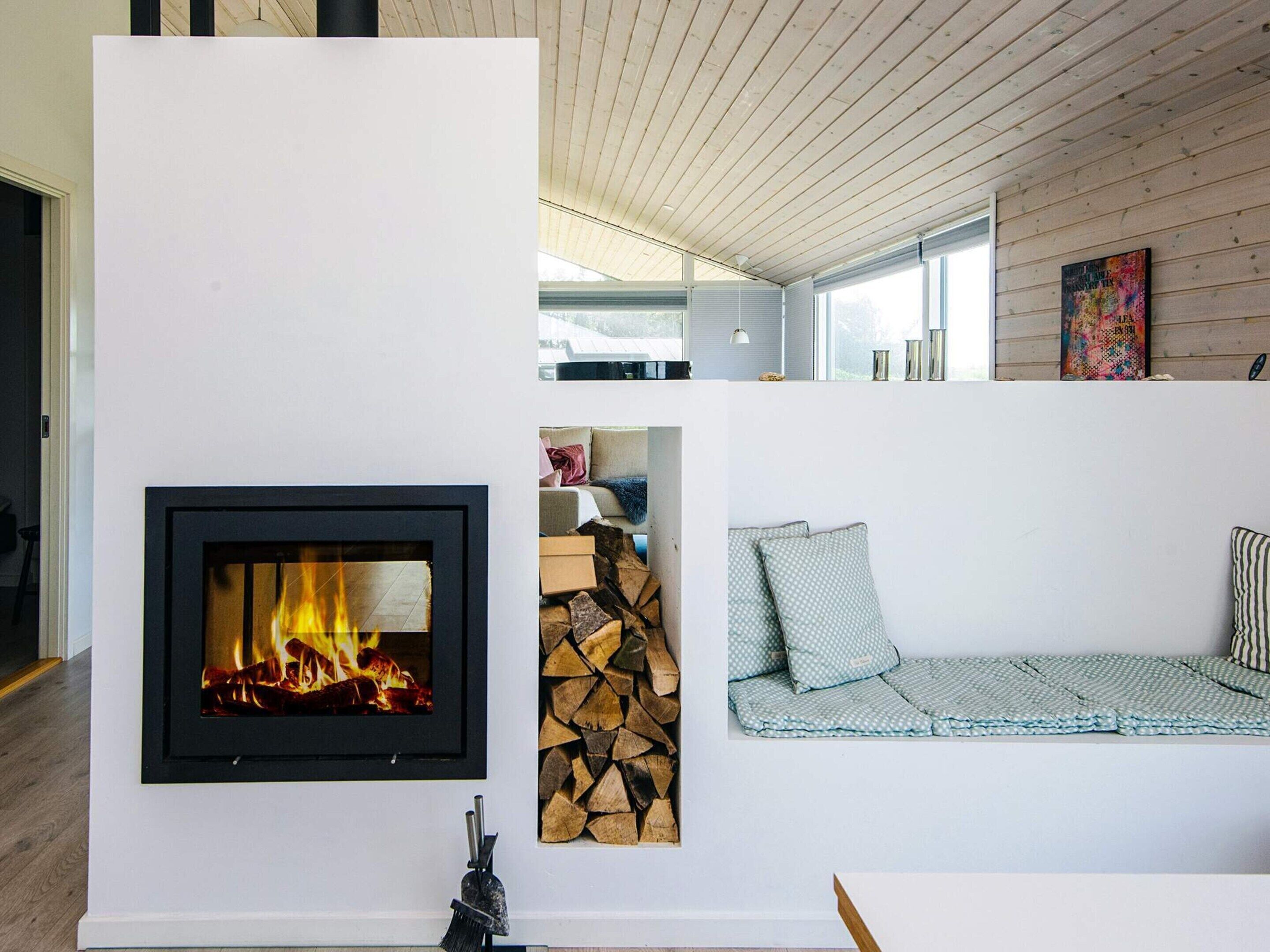 A modern living room with a fireplace, a stack of firewood, and a sofa with patterned cushions.