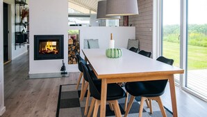 Un espace repas moderne doté d’une cheminée, d’un plafond en bois et de grandes fenêtres offrant une vue sur l’extérieur.