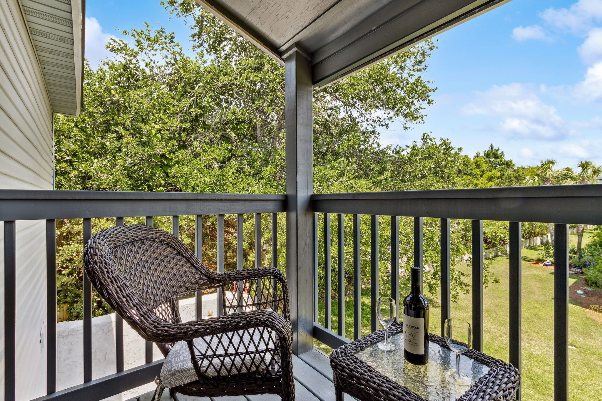 Un espace extérieur aménagé, abrité et doté de chaises et d’une table en osier, offrant une vue sur un jardin arboré et un ciel dégagé.