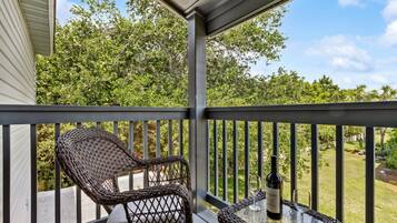 A covered outdoor seating area with wicker chairs and a table, overlooking a garden with trees and a clear sky.