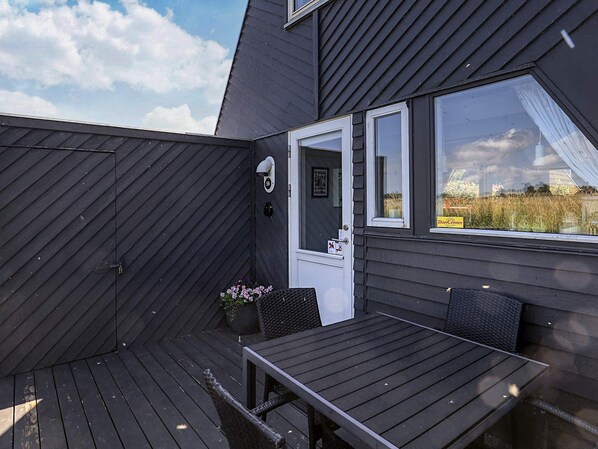 A dark wooden deck with a table and chairs, a door with a number sign, and a window with a curtain.