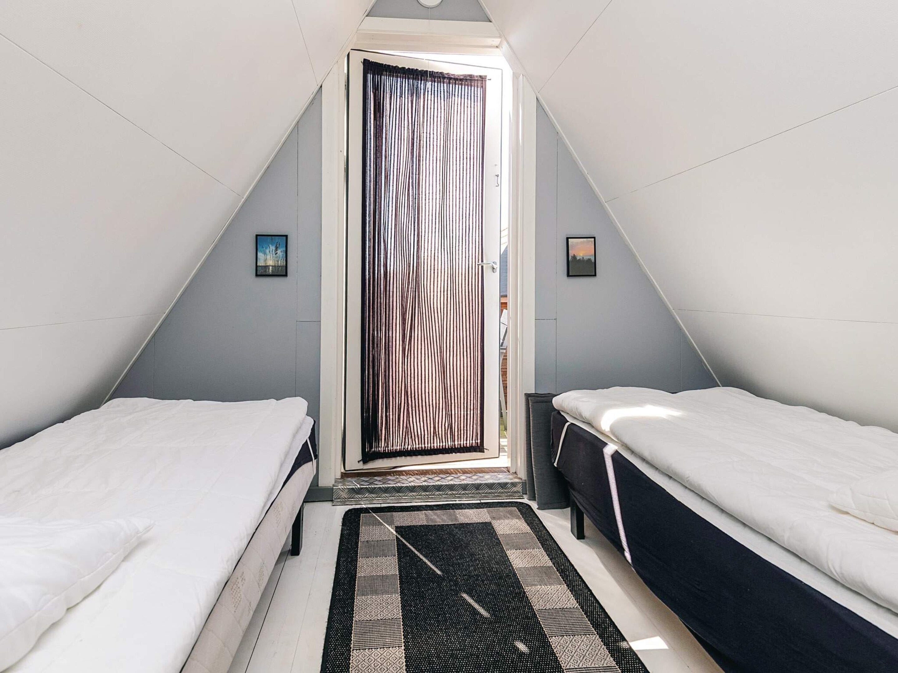 A small, triangular room with two beds, a window with curtains, and a door with a striped pattern.