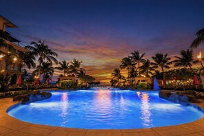A large outdoor swimming pool with a sunset view, surrounded by palm trees and lounge areas.