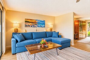 Una sala de estar con un sofá azul, una mesa de centro de vidrio y un cuadro que representa una playa.