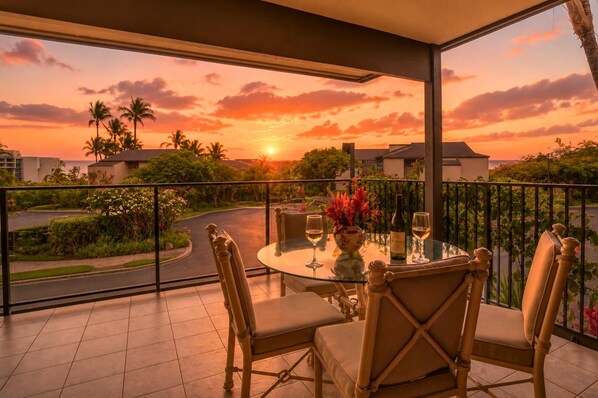 Een balkon met een tafeltje voor twee, met uitzicht op een zonsondergang met palmbomen en een weg.