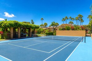 An outdoor tennis court with a blue surface, surrounded by greenery and palm trees.