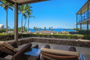 A covered outdoor seating area with wicker furniture, a stone wall, and a view of the ocean and palm trees.