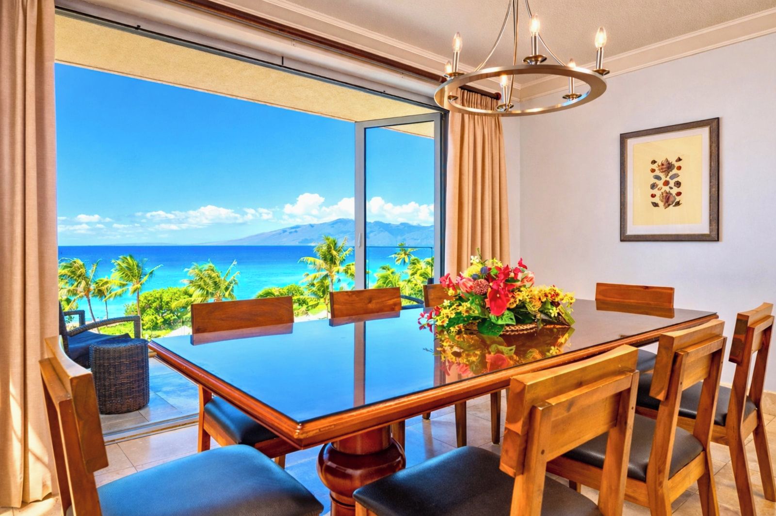 Un coin repas avec une table en verre, des chaises en bois, un lustre et une vue sur l’océan.