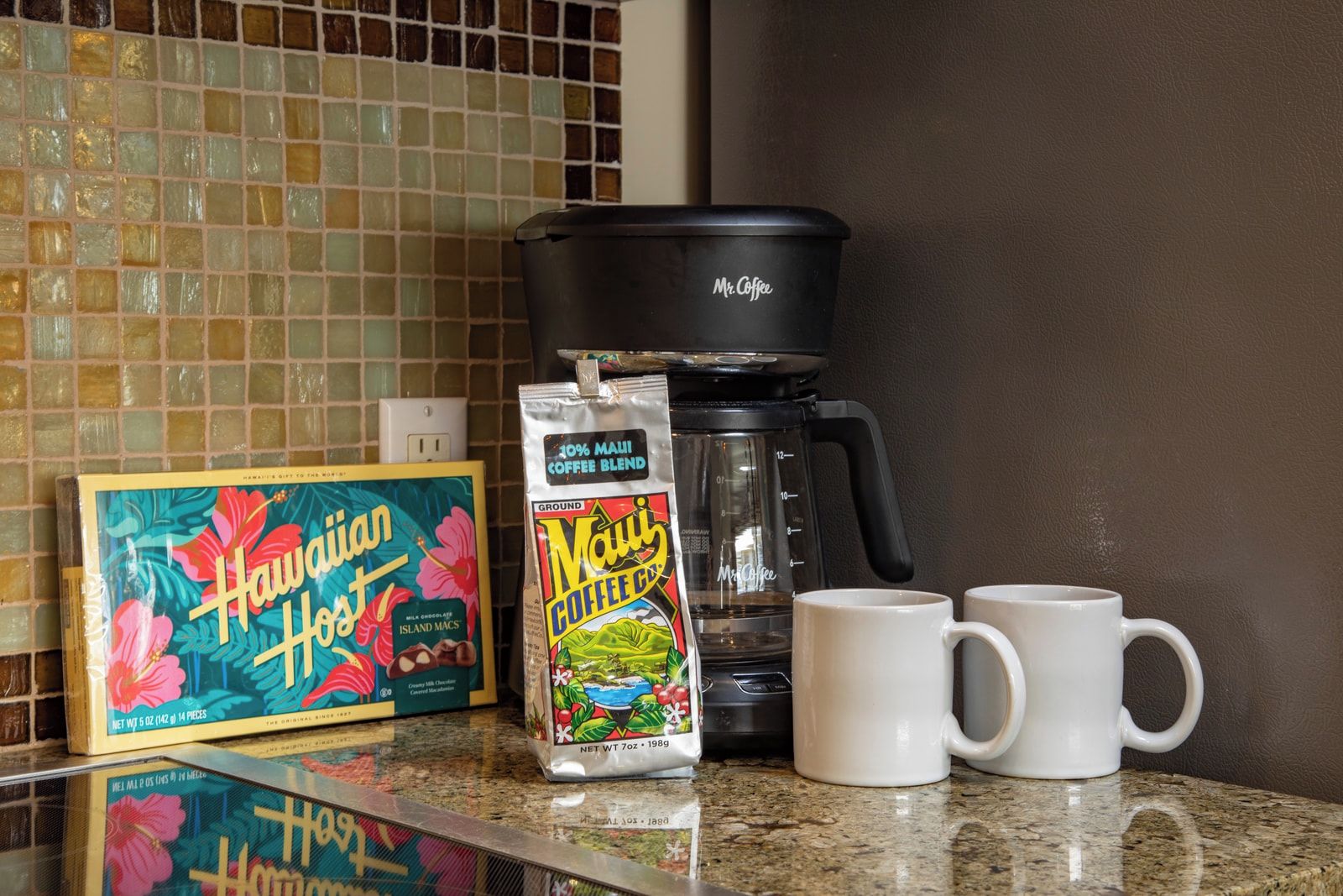 Een keukenblad met een koffiezetapparaat, een doos Hawaiian Hot Island Macas, een zak Maui Coffee blend, en twee witte mokken.